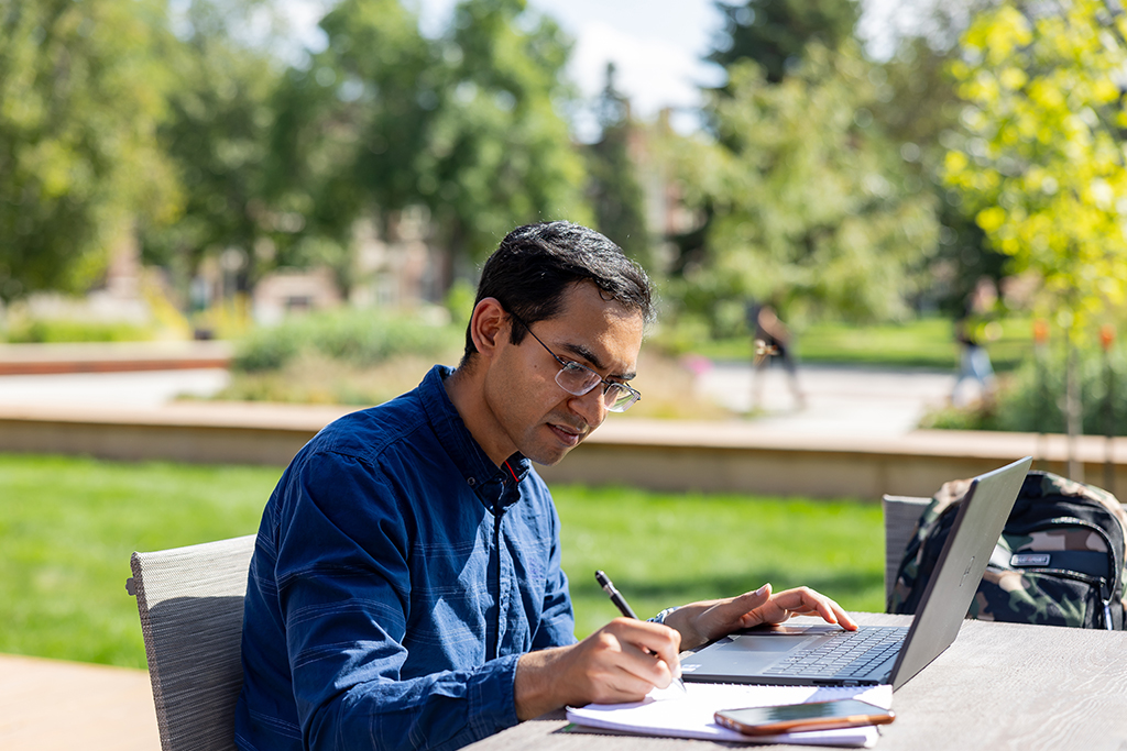 a student sits outdoors at a table, working on a laptop and writing in a notebook, with trees and blurred figures in the background