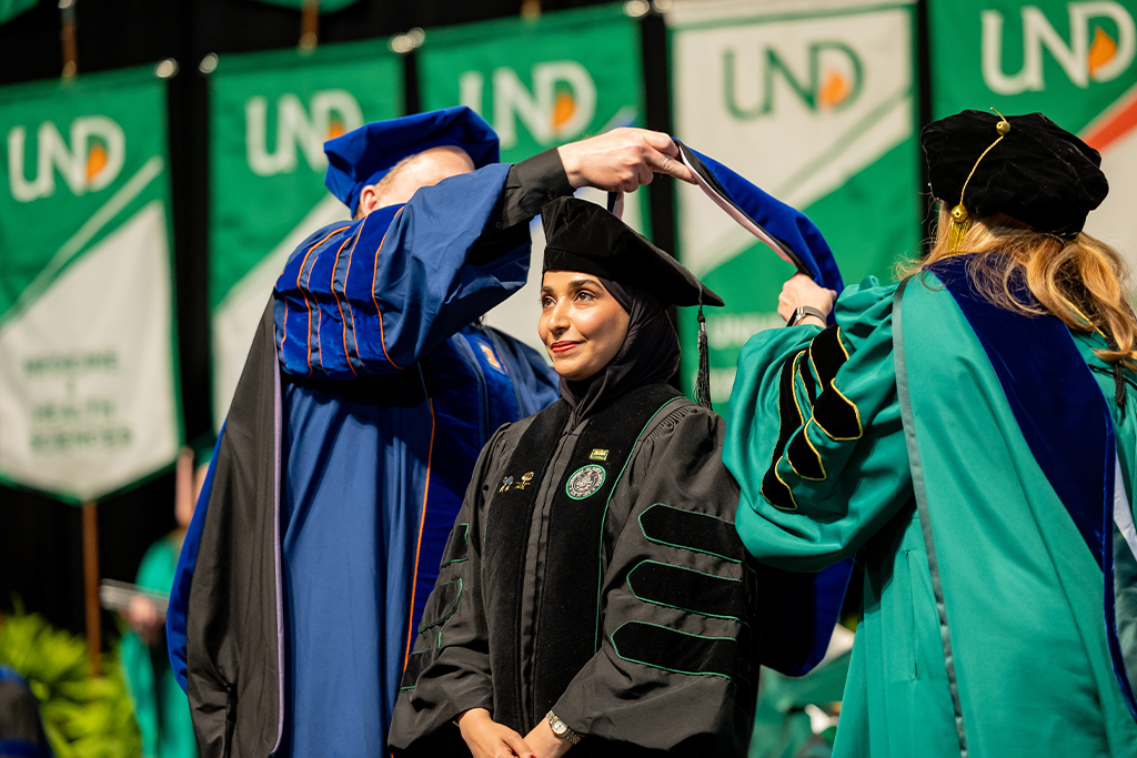 at a graduation ceremony, an individual is hooded by two faculty members in academic regalia—one in a blue gown and the other in green—with 'UND' and 'WelcomD Family' banners e to the UNin the background