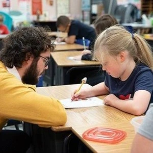 A teacher talking to a young student across her desk.