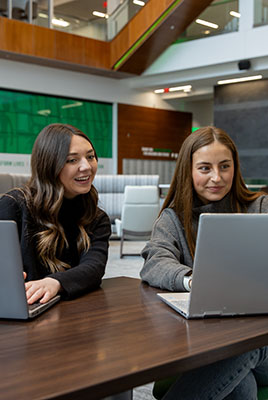 Two students sitting at a table with their laptops. Student on the right is looking over the sholder of the student on the left. Both students are smiling.