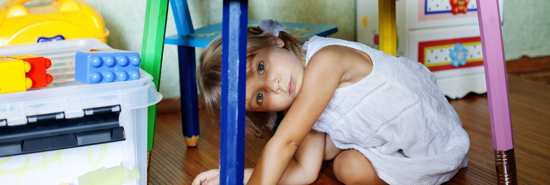 A young girl playing underneath a classroom table, looking sideways at the camera.