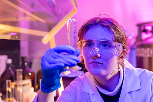 Student wearing eye protection looks at a beaker full of water in a lab.